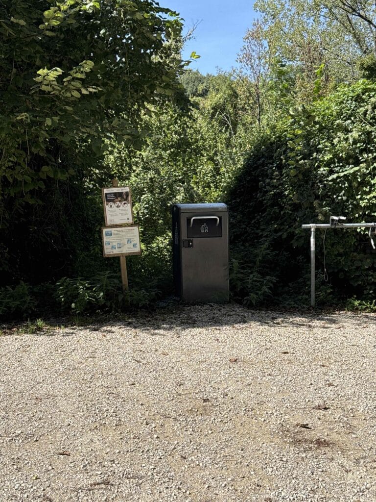 Conteneur poubelle et panneaux près de buissons verts.